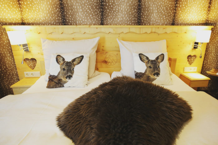 Holzdoppelbett mit weißer Bettwäsche, braunem Tierfell in der Mitte, zwei Kissen mit Rehköpfen und brauner Rehfell-Tapete an der Wand