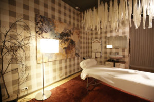 Seitenansicht weißer Massageliege, weißer Dekostoff mit Lichterkette hängt von der Decke, warm leuchtende Stehlampe links