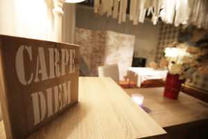 Holzschild mit weißer Aufschrift "Carpe Diem", im Hintergrund ist verschwommen eine Massageliege zu sehen