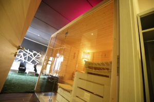 Bild einer Sauna mit Glasfront in einem gemütlichen Raum, weiße Liegen und Kunstrasen im Hintergrund