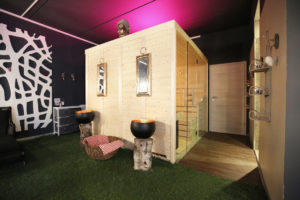 Außenansicht einer Holzsauna in einem schwarzen Raum mit Kunstrasen und ansprechender Dekoration