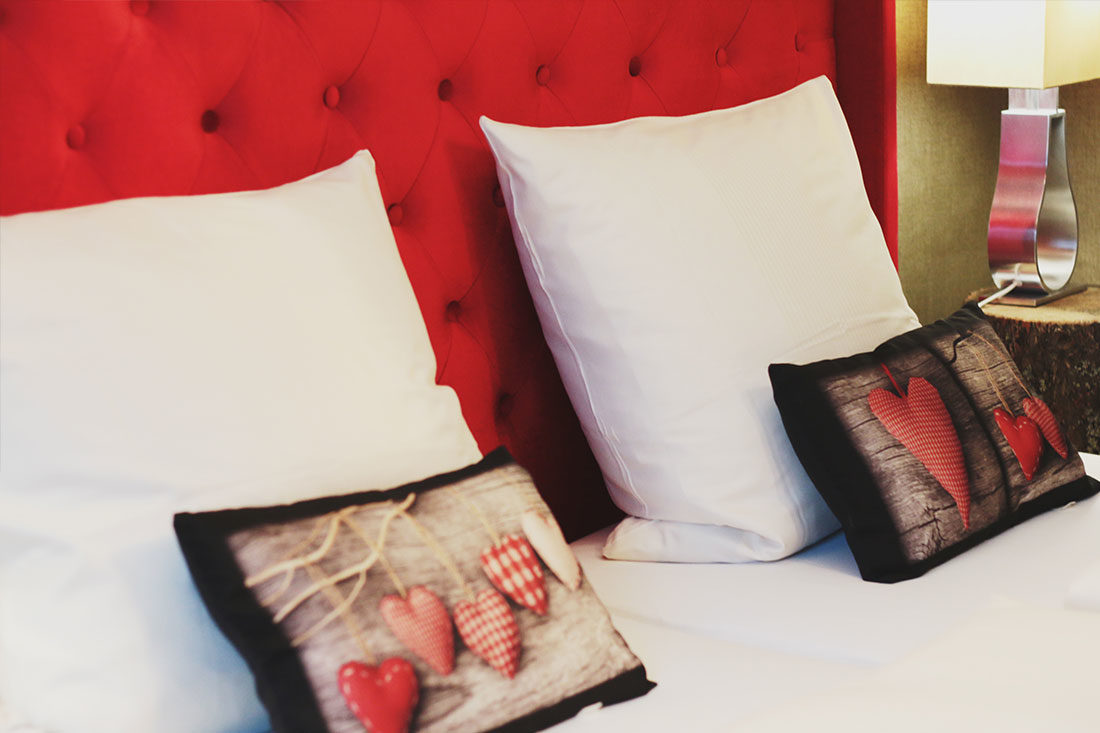 Rotes Doppelbett mit weißer Bettwäsche, zwei kleine schwarze Kissen mit roten Herzen vor zwei großen weißen Kissen