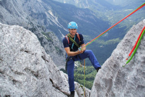 Ein Mann grinst während er durch Seile gesichert ist und auf einem hohen Berg steht, im Hintergrund ein Wald und eine Bergregion zu sehen