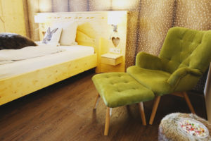 Holzbett, braune Rehfelltapete an der Wand, gemütlicher grüner Sessel mit Hocker
