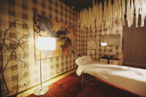 Seitenansicht weißer Massageliege, weißer Dekostoff mit Lichterkette hängt von der Decke, warm leuchtende Stehlampe links