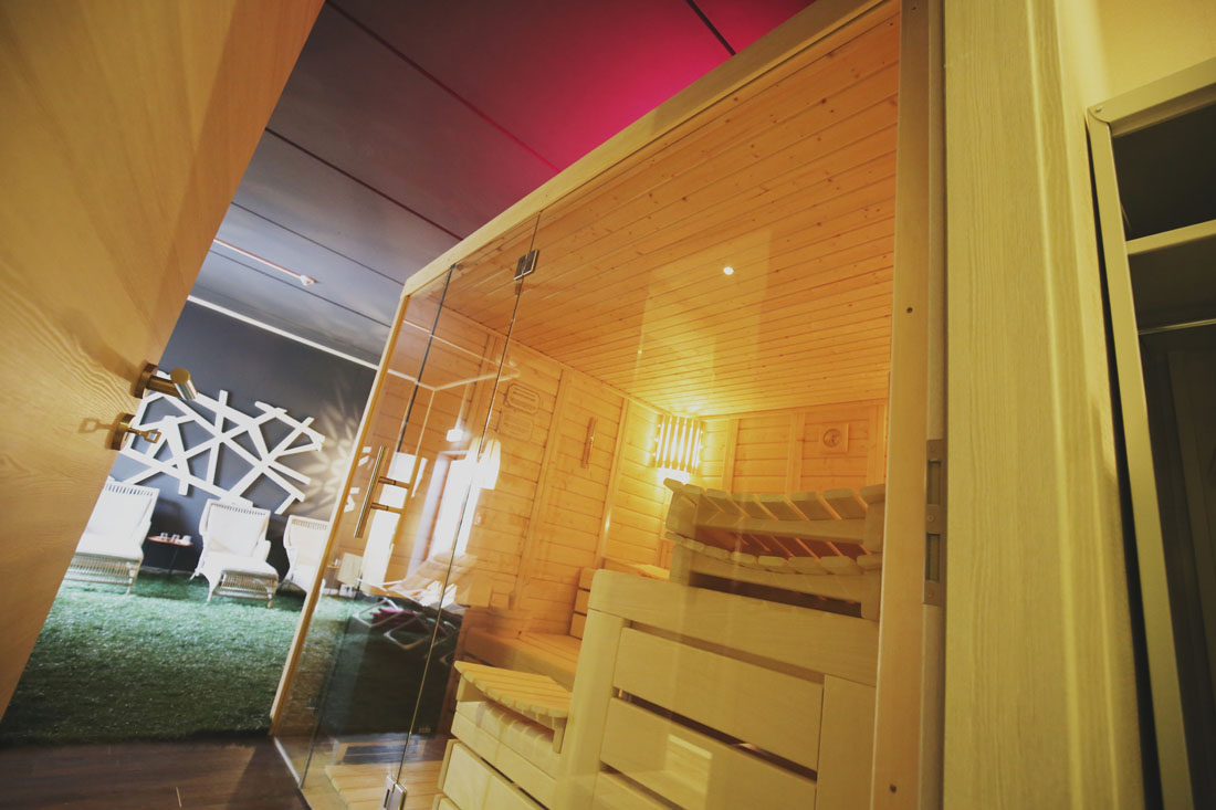 Bild einer Sauna mit Glasfront in einem gemütlichen Raum, weiße Liegen und Kunstrasen im Hintergrund