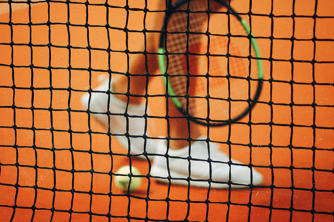 Schwarzes Tennisnetz von der Nähe, im Hintergrund eine Person mit einem Tennisschläger und weißen Schuhen, Tennisball auf dem Boden